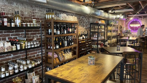 La imagen muestra un ambiente elegante y acogedor, que parece una vinoteca o un bistró gourmet. Las paredes de ladrillo visto y el techo con vigas de madera y caños metálicos vistos dan un toque industrial y rústico al espacio. Descripción del entorno: Estanterías de madera y metal repletas de productos artesanales italianos, entre ellos botellas de vino, pastas, conservas, salsas, aceites y legumbres. Mesas y sillas de madera oscura con detalles en hierro, que crean un ambiente cálido y sofisticado, ideal para degustaciones o reuniones de convivencia. Una iluminación tenue, con luces cálidas y un sugerente letrero de neón de color violeta al fondo con la inscripción “Visionarium Buon Appetito”, que aporta un toque moderno y distintivo al recinto. Un rincón con una persona que parece preparar o presentar productos, lo que sugiere que puede ser un lugar dedicado al descubrimiento y promoción de la excelencia enogastronómica italiana. La combinación de estos elementos transmite una fuerte referencia a la tradición italiana, con especial atención a la calidad de los productos y a la estética acogedora, perfecta para quienes buscan una auténtica experiencia de sabores y cultura.