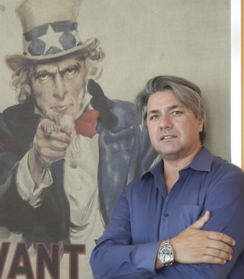 Paolo Odierna in posa davanti al celebre poster di Uncle Sam 'I Want You' - L'immagine mostra un uomo con camicia blu e orologio al polso, in piedi con le braccia incrociate, davanti al famoso manifesto di reclutamento statunitense. Questa scena simboleggia il legame con il business negli USA e l'opportunità per imprenditori italiani di espandersi nel mercato americano. Us Company Advisor offre consulenza strategica per aziende che vogliono crescere negli Stati Uniti.