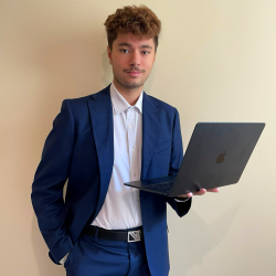 Tommaso Odierna - Social Media Manager at Us Company Advisor - The image shows Tommaso Odierna, a young professional in a blue suit with a white shirt, a branded belt and an Apple laptop in his hand, symbolizing his role in digital marketing. With a confident and dynamic expression, he represents the strategic management of social media. Us Company Advisor helps Italian companies develop an effective digital presence for the US market.