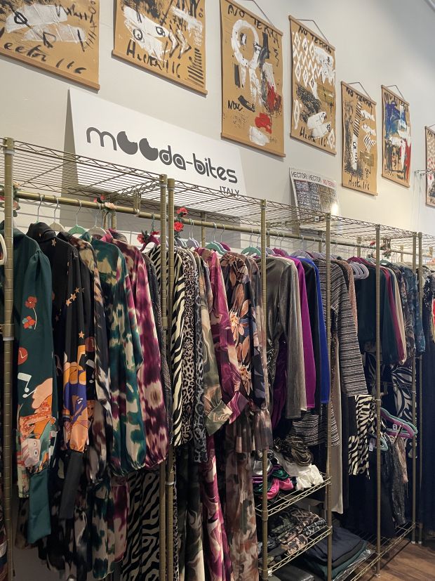 La imagen muestra una boutique o showroom de moda, con una muestra de prendas con diseños originales y estampados llamativos. El ambiente de la tienda se caracteriza por un estilo artístico y contemporáneo, con detalles creativos y refinados. Descripción del entorno: Ropa colorida y actual, con estampados florales, animales y abstractos, expuesta en perchas doradas de diseño minimalista. Mobiliario con un toque industrial, con estanterías de metal dorado y una ordenada disposición de la ropa. Arte en las paredes, con obras abstractas sobre cartón crudo, que añaden un elemento artístico y alternativo al concepto de la tienda. Un cartel con el logo "Mood da-bites Italy" sugiere una marca italiana centrada en diseños únicos y refinados. Pequeños detalles decorativos, como flores colocadas entre las prendas, contribuyen a crear un ambiente acogedor y sofisticado. Esta boutique parece especializada en moda elegante y creativa, probablemente una marca emergente o una muestra de prendas de diseño artesanales y Made in Italy.