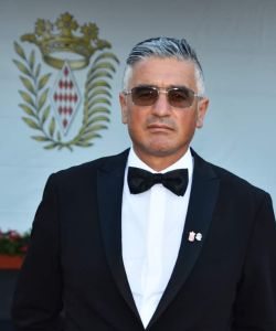 Giosuè Gilberto Di Molfetta - Manager of Us Company Advisor - Professional portrait of Giosuè Gilberto Di Molfetta with salt and pepper hair, sunglasses and black tuxedo with bow tie, in an elegant setting with a noble coat of arms in the background. The exclusive atmosphere and sophisticated look convey authority and prestige. Us Company Advisor offers high-level consultancy for Italian companies that want to expand into the US market.