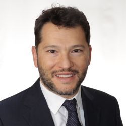 Alessandro Betro - Gerente de Asesor de la Compañía de EE. UU. - Retrato profesional de un hombre con cabello rizado y barba corta, vestido con chaqueta oscura, camisa blanca y corbata de lunares, sobre un fondo blanco. Su sonrisa transmite profesionalidad y seriedad. Us Company Advisor ofrece consultoría estratégica para empresarios italianos que desean expandir sus negocios en los Estados Unidos.
