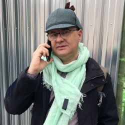 Stefano Palai - Manager of Us Company Advisor - The image shows Stefano Palai with glasses and a Sherlock Holmes-style hat, wrapped in a light green scarf, while talking on the phone. He wears a dark jacket and carries a backpack, suggesting a dynamic and professional context. Us Company Advisor offers strategic consulting for Italian companies that want to expand in the United States, with a personalized and innovative approach.