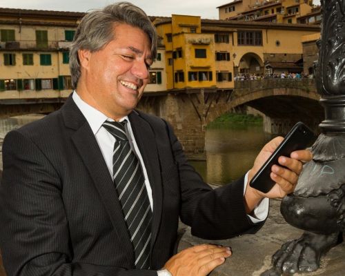 L'immagine mostra tre uomini in posa davanti a uno schermo su cui è proiettato il logo "NEXXT PRESENTA", caratterizzato da colori che richiamano il tricolore italiano. L'uomo a sinistra indossa un abito scuro con camicia bianca, quello al centro un blazer beige con camicia bianca, mentre quello a destra veste un maglione nero con motivi geometrici e una sciarpa. Sullo sfondo è visibile un banner verticale con la scritta "Confartigianato", suggerendo un evento legato all'imprenditoria o al commercio. L'ambiente appare come una sala conferenze o un luogo di presentazione formale.