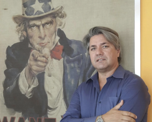 L'immagine mostra tre uomini in posa davanti a uno schermo su cui è proiettato il logo "NEXXT PRESENTA", caratterizzato da colori che richiamano il tricolore italiano. L'uomo a sinistra indossa un abito scuro con camicia bianca, quello al centro un blazer beige con camicia bianca, mentre quello a destra veste un maglione nero con motivi geometrici e una sciarpa. Sullo sfondo è visibile un banner verticale con la scritta "Confartigianato", suggerendo un evento legato all'imprenditoria o al commercio. L'ambiente appare come una sala conferenze o un luogo di presentazione formale.
