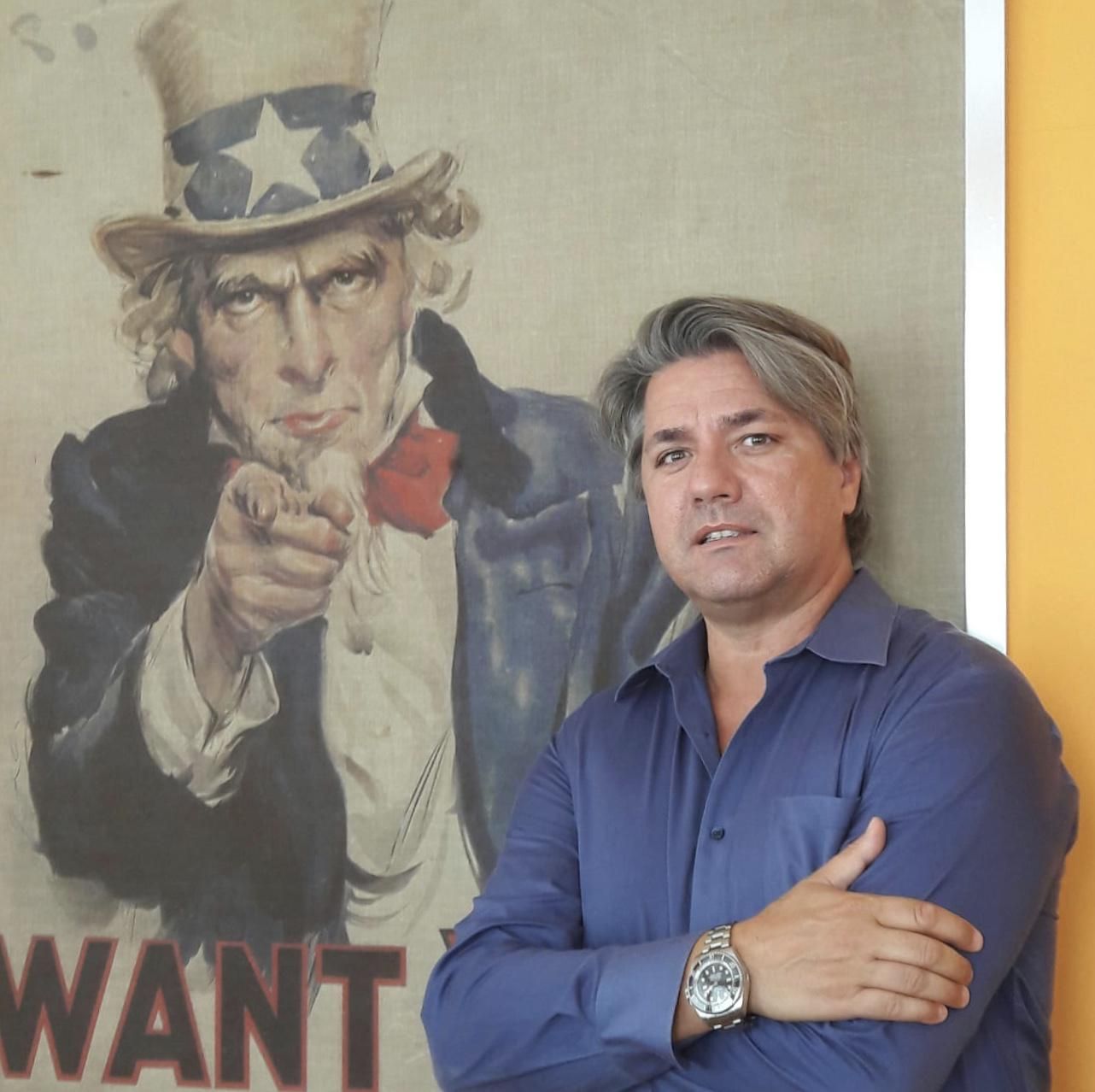 Paolo Odierna in posa davanti al celebre poster di Uncle Sam 'I Want You' - L'immagine mostra un uomo con camicia blu e orologio al polso, in piedi con le braccia incrociate, davanti al famoso manifesto di reclutamento statunitense. Questa scena simboleggia il legame con il business negli USA e l'opportunità per imprenditori italiani di espandersi nel mercato americano. Us Company Advisor offre consulenza strategica per aziende che vogliono crescere negli Stati Uniti.