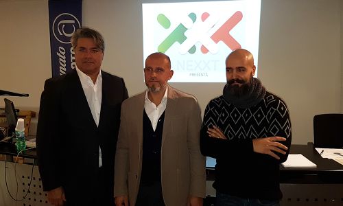 L'immagine mostra tre uomini in posa davanti a uno schermo su cui è proiettato il logo "NEXXT PRESENTA", caratterizzato da colori che richiamano il tricolore italiano. L'uomo a sinistra indossa un abito scuro con camicia bianca, quello al centro un blazer beige con camicia bianca, mentre quello a destra veste un maglione nero con motivi geometrici e una sciarpa. Sullo sfondo è visibile un banner verticale con la scritta "Confartigianato", suggerendo un evento legato all'imprenditoria o al commercio. L'ambiente appare come una sala conferenze o un luogo di presentazione formale.