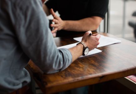 Press Office and Corporate Communication Strategy - The image shows two people engaged in a discussion on official documents, sitting at a wooden table with papers and pen in hand. The context represents the planning of press releases and media relations strategies. Us Company Advisor helps Italian companies gain visibility in the US market through professional management of communications and public relations.