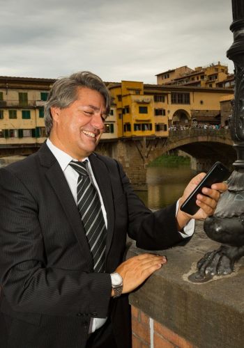 L'immagine mostra Paolo Odierna con capelli grigi pettinati all'indietro, vestito con un elegante completo scuro a righe sottili, una camicia bianca e una cravatta a righe diagonali. Sorride mentre guarda lo schermo del suo smartphone, che tiene con una mano. L'altra mano è appoggiata su un muretto accanto a un lampione decorativo. Sullo sfondo, si distingue il Ponte Vecchio di Firenze con le sue caratteristiche botteghe e numerosi turisti che passeggiano sopra di esso. Il cielo è coperto, ma la scena è ben illuminata, probabilmente con l'ausilio di un flash fotografico.