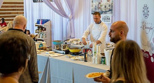 L'immagine mostra un evento gastronomico in cui uno chef, vestito con una giacca da cuoco bianca, sta preparando dei piatti su un bancone coperto da una tovaglia chiara. Davanti a lui ci sono padelle, pentole, bottiglie di condimenti e altri ingredienti. Il pubblico, composto da diverse persone, osserva attentamente la preparazione. Un uomo con la testa rasata tiene in mano un piatto con del cibo, mentre una donna accanto a lui guarda interessata. Sullo sfondo si notano decorazioni eleganti con tende bianche e banner promozionali di aziende alimentari, suggerendo che l'evento potrebbe essere una degustazione o una dimostrazione culinaria in un contesto fieristico.