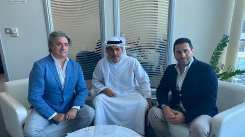L'immagine mostra tre uomini seduti su poltrone bianche in un ambiente moderno e professionale, probabilmente un ufficio o una sala riunioni. L'uomo a sinistra indossa un abito blu chiaro con una camicia bianca, ha capelli grigi e un aspetto distinto. L'uomo al centro è vestito con un tradizionale abito bianco arabo (kandura) e un copricapo (ghutra), con un'espressione seria e attenta. L'uomo a destra indossa un blazer blu scuro con una camicia bianca e pantaloni chiari, con una postura rilassata e un sorriso accennato. Sul tavolino davanti a loro ci sono alcuni documenti, suggerendo un incontro di lavoro o una trattativa. Sullo sfondo si vedono grandi finestre con vetri e pannelli a righe, lasciando entrare molta luce naturale.