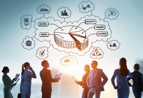 Un gruppo di professionisti d'affari in controluce discute strategie aziendali davanti a un grafico concettuale digitale. Il grafico include una rappresentazione a torta con parole chiave come 'Global', 'Quality', 'Design', 'Slogan' e 'Media', collegate da nuvole concettuali. L'immagine evoca innovazione, networking e sviluppo aziendale internazionale.