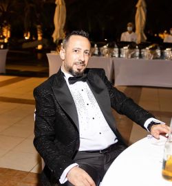 Hussam Abdulkhadir Al Hamad - Manager di Us Company Advisor - L'immagine mostra Hussam Abdulkhadir Al Hamad elegante con barba corta, vestito con uno smoking nero con dettagli brillanti, camicia bianca e papillon, seduto a un tavolo durante un evento di gala. Lo sfondo con camerieri e tavoli imbanditi suggerisce un contesto esclusivo e professionale. Us Company Advisor fornisce consulenza strategica per aziende italiane che desiderano espandersi nel mercato statunitense.