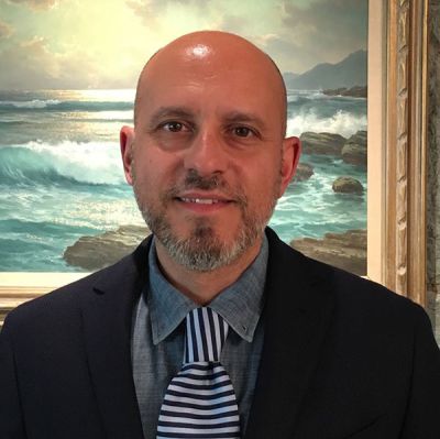 Lorenzo Lisi in abito elegante con cravatta a righe davanti a un dipinto marino - L'immagine mostra Lorenzo Lisi con barba corta e sorriso, vestito in modo formale con giacca scura e cravatta a righe, posizionato davanti a un quadro raffigurante un paesaggio marino con onde e cielo al tramonto. Us Company Advisor offre consulenza personalizzata per imprenditori italiani che vogliono espandersi nel mercato statunitense.