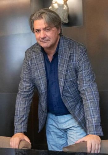 L'immagine mostra Paolo Odierna con capelli grigi pettinati all'indietro, vestito con una giacca a quadri blu e grigia, una camicia blu scuro e pantaloni chiari. È in una posizione leggermente inclinata in avanti, con le mani appoggiate su un tavolo o una superficie. Lo sfondo presenta un ambiente elegante e moderno, con pareti scure e una scultura bianca illuminata in alto, che aggiunge un tocco artistico alla scena. L'uomo ha un'espressione sicura e un leggero sorriso.