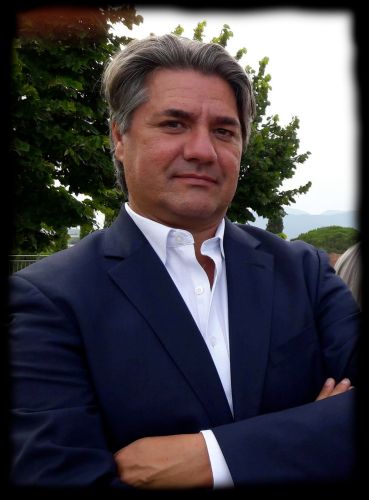 L'immagine mostra Paolo Odierna con capelli grigi ben curati, vestito con un elegante abito blu scuro e una camicia bianca senza cravatta. Ha un'espressione seria e determinata, con le braccia incrociate in una posa sicura. Lo sfondo è all'aperto, con alberi verdi e una vista sulle montagne in lontananza. L'immagine ha un effetto vignettatura nera sui bordi, che attira l'attenzione sul soggetto principale.