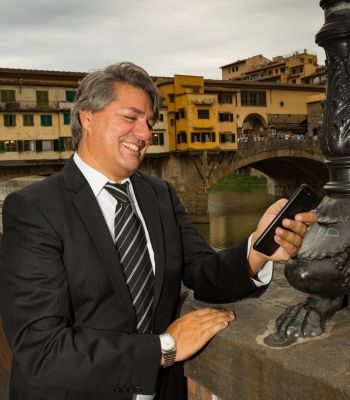 L'immagine mostra Paolo Odierna con capelli grigi pettinati all'indietro, vestito con un elegante completo scuro a righe sottili, una camicia bianca e una cravatta a righe diagonali. Sorride mentre guarda lo schermo del suo smartphone, che tiene con una mano. L'altra mano è appoggiata su un muretto accanto a un lampione decorativo. Sullo sfondo, si distingue il Ponte Vecchio di Firenze con le sue caratteristiche botteghe e numerosi turisti che passeggiano sopra di esso. Il cielo è coperto, ma la scena è ben illuminata, probabilmente con l'ausilio di un flash fotografico.